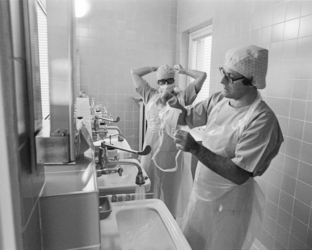Photo des lavabos de la salle opératoire, 2 chirurgiens se lavent les mains pour se préparer à une opération (Docteur Guy Mandres, Docteur Norbert Weydert) . Ils portent des vêtements spéciaux, portés uniquement dans le cadre opératoire pour éviter les infections. Leurs cheveux et leur région naso-buccale sont aussi couverts. Entretemps le patient est mis sur la table d’opération et mis sous anesthésie. Le respect des mesures préopératoires et un bon travail d’équipe sont très importants pendant cette phase afin de réduire le temps d’anesthésie à un minimum.
A l’époque, il y avait 3 salles opératoires à la clinique Ste Zithe, où on faisait des interventions chirurgicales à l’abdomen, au thorax et aux os dans des conditions d'aseptie optimales.
A coté il y avait des dépôts pour le matériel stérile, un très grand stérilisateur dans les pièces de conditionnement de matériel et d’équipements, des salles de permanence pour les collaborateurs.